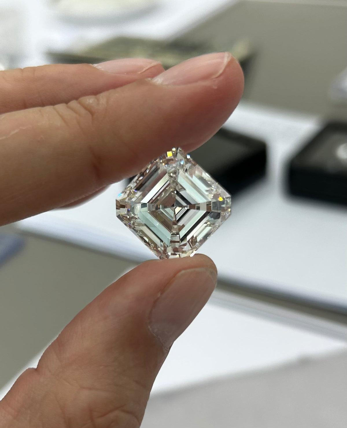 Main de Arnaud Pellegrin tenant entre le pouce et l’index un diamant transparent de taille carrée (type Asscher ou princesse), orienté en losange, avec des facettes géométriques bien visibles et des reflets lumineux.