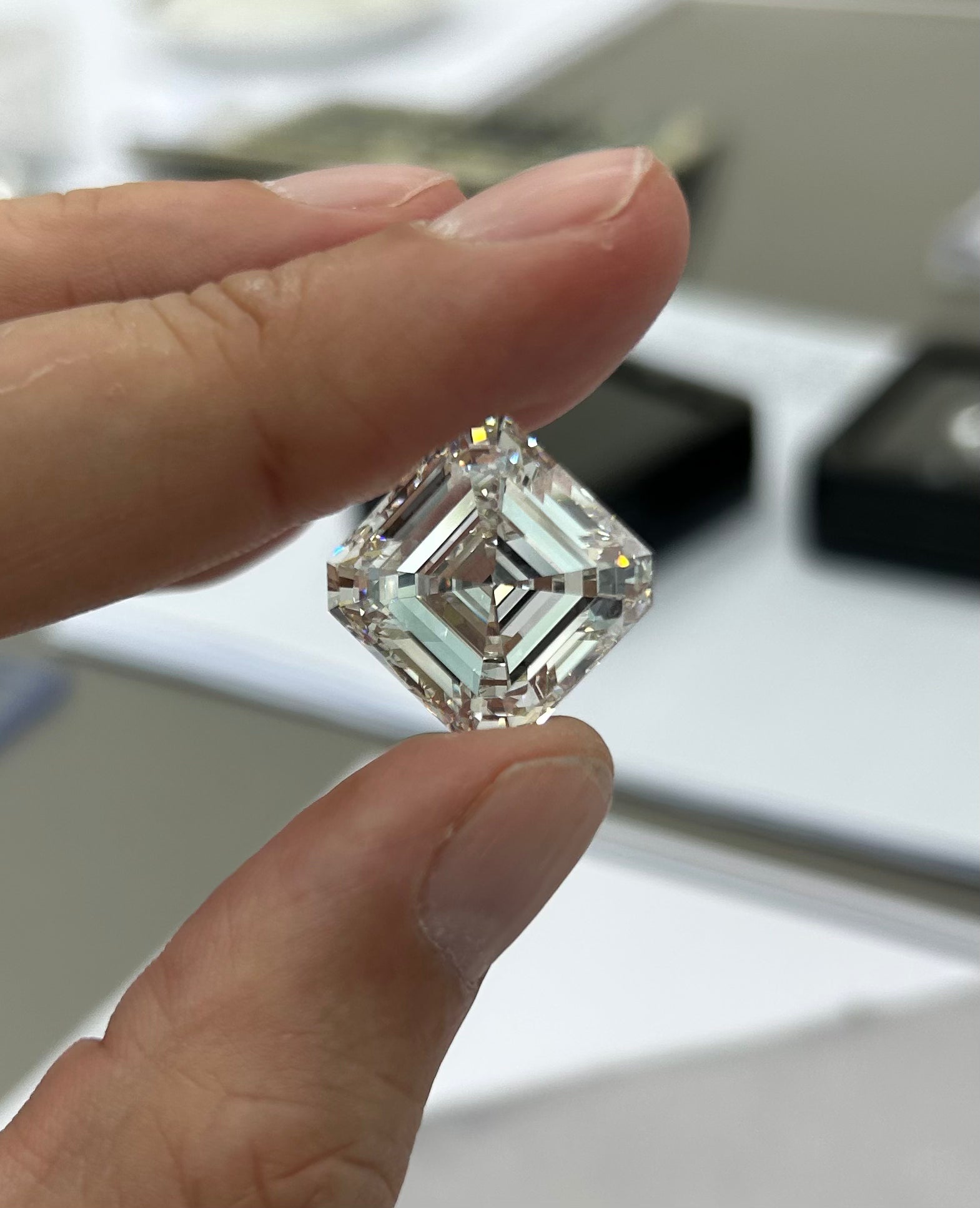 Main de Arnaud Pellegrin tenant entre le pouce et l’index un diamant transparent de taille carrée (type Asscher ou princesse), orienté en losange, avec des facettes géométriques bien visibles et des reflets lumineux.