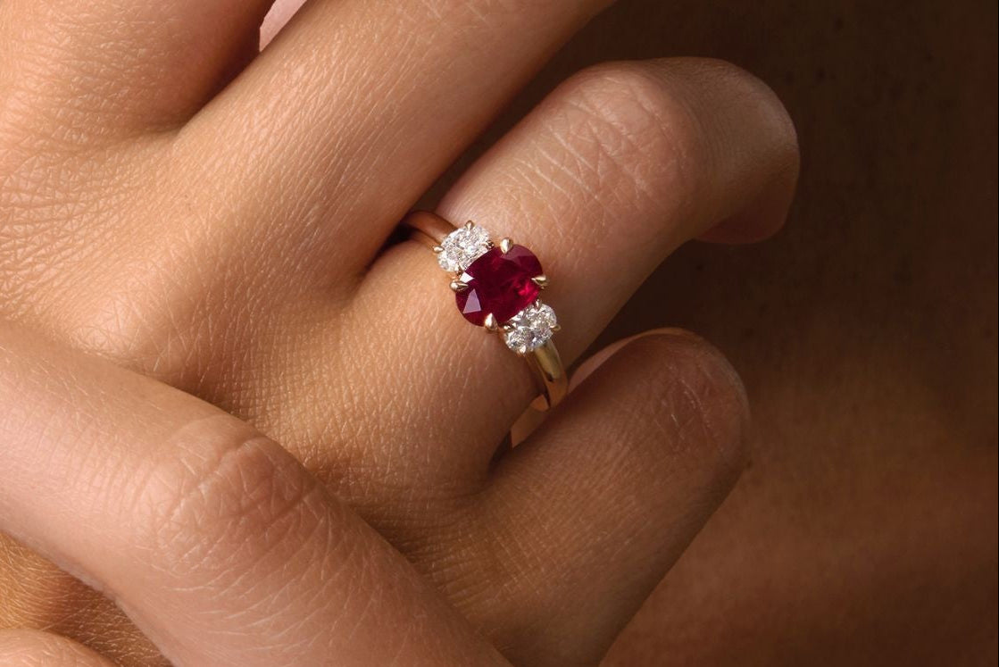 Bague Trilogie en or blanc sertie d’un saphir bleu ovale entouré de deux diamants, portée à la main, et bague Trilogie en or jaune ornée d’un rubis rouge taille coussin encadré de diamants, vue en gros plan sur peau hâlée.