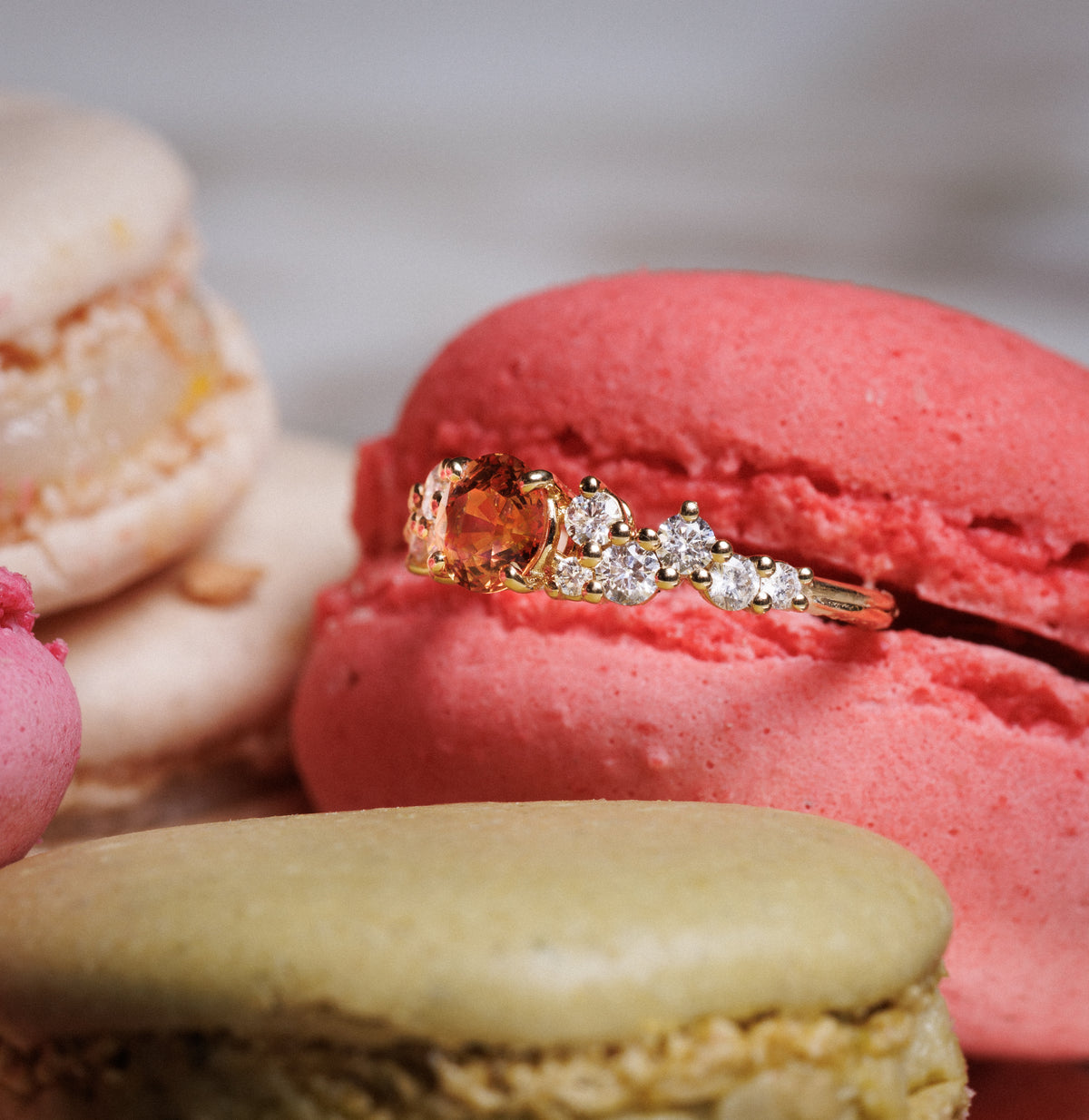 Bague en or jaune sertie d’un saphir orange et de diamants, posée sur un macaron rose