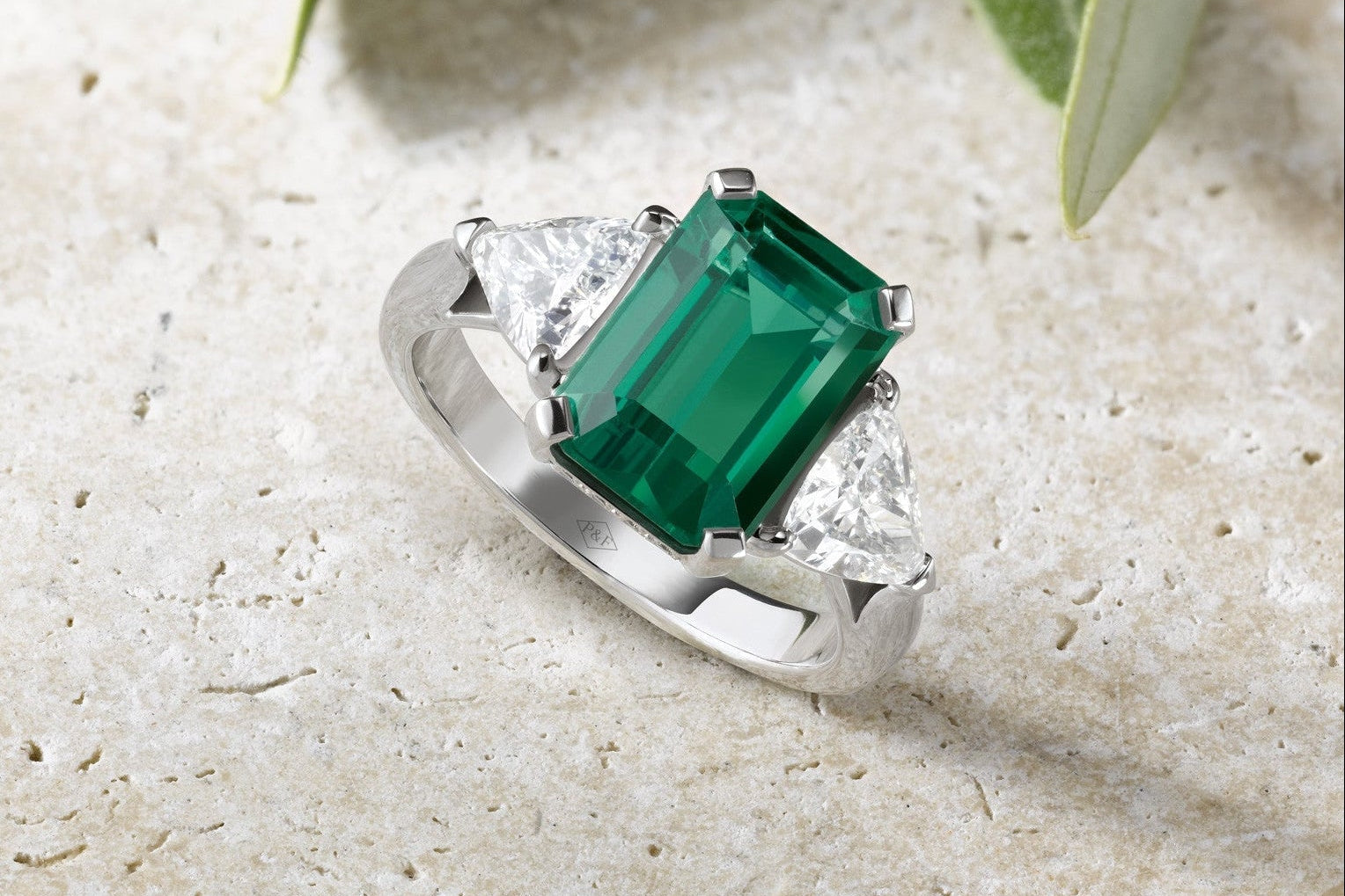 Bague Trilogie en or blanc sertie d’une émeraude taille émeraude encadrée de deux diamants taille trillion, posée sur une pierre claire avec feuilles vertes en arrière-plan.