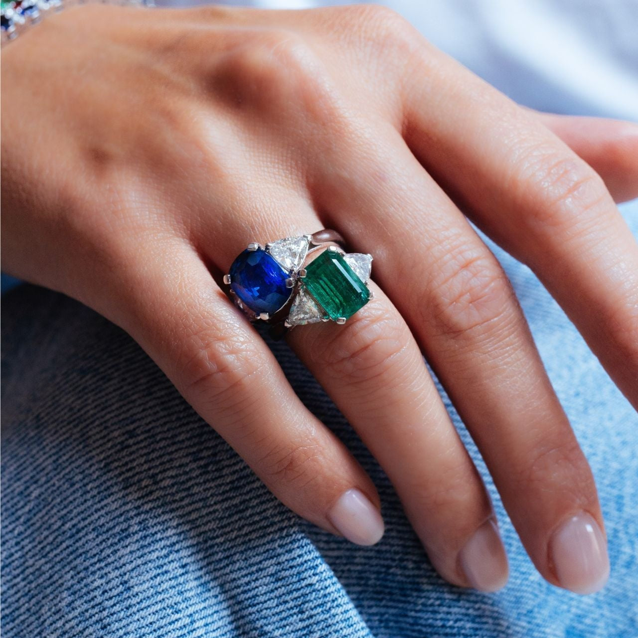 Bague Trilogie en or blanc portée à la main, composée d’un saphir bleu ovale et d’une émeraude taille émeraude, encadrés de diamants taille triangle, sur fond de denim clair.